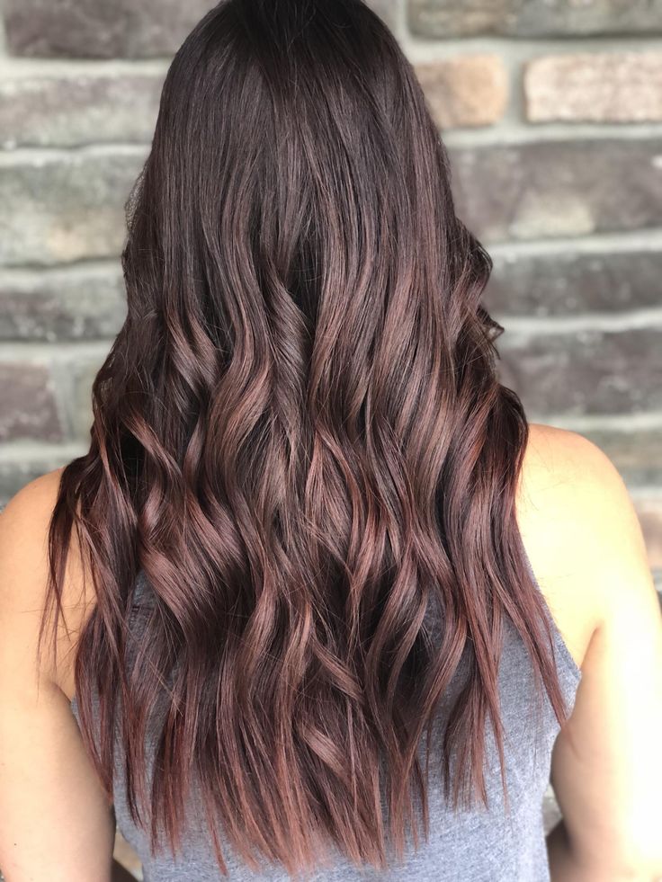 Back view of a person with long, dark brown hair in loose burgundy-tinted waves against a brick wall backdrop, wearing a gray tank top.